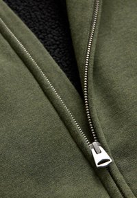 Felpa con zip di colore verde oliva, caratterizzata da una fodera in fleece nera di contrasto, con zip metallica e tessuto morbido e testurizzato.