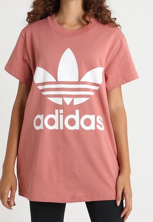 Kobieta w luźnej, różowej koszulce Adidas z krótkim rękawem i dużym białym logo oraz czarnych spodniach.