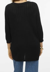 Nainen, jolla on päällään löysä musta neulepusero, jonka hihat ovat kolme neljäsosaa, sekä siniset farkut, nähtynä takaa. Hänen vasemmassa ranteessaan on kultainen rannekoru.