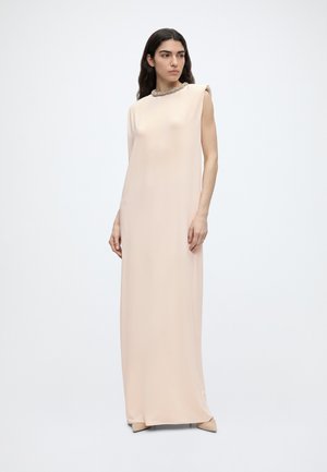 Frau mit langen dunklen Haaren, die ein ärmelloses, bodenlanges beiges Kleid mit verzierter Halslinie trägt, steht vor einem schlichten weißen Hintergrund.