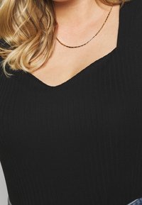 Svart ribbad bodysuit med V-ringning, som har en mjuk textur och åtsittande siluett. En guldhalsband med en subtil glans syns.