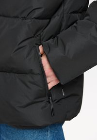 Doudoune noire avec une poche latérale zippée, présentant une texture douce et matelassée ainsi que des poignets élastiqués, présentée à côté de jeans en denim bleu.