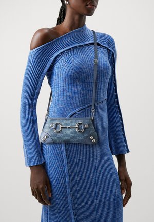 Femme portant une robe bleue côtelée à épaules dénudées, portant un petit sac bandoulière bleu avec des détails en métal sur la boucle.