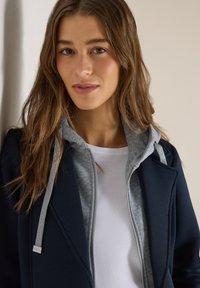 Jeune femme aux longs cheveux bruns portant un manteau bleu marine par-dessus un sweat à capuche gris zippé et une chemise blanche, debout contre un mur de couleur claire.