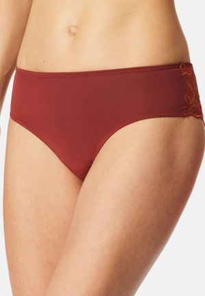 Žena nosící hipsterské kalhotky rezavé barvy s krajkovými detaily po stranách na bílém pozadí.