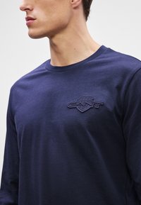 Mornarsko dolgimanžetni T-shirt iz mehkega bombaža z okroglim ovratnikom. Ima dvignjen GANT logo v usklajeni barvi na prsih.