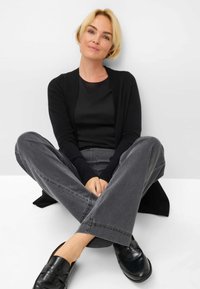Zwarte cardigan over een aansluitend zwart topje, gecombineerd met grijze wijde broekspijpen en zwarte instapschoenen, zittend met gekruiste benen op een witte ondergrond.