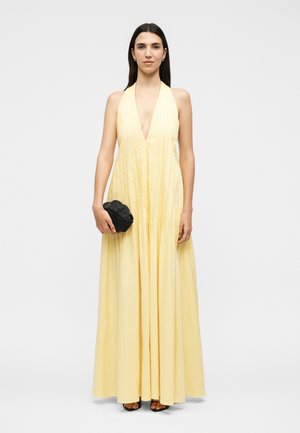 Femme en robe maxi sans manches jaune pâle à col halter, tenant une petite pochette noire, debout devant un fond clair uni.