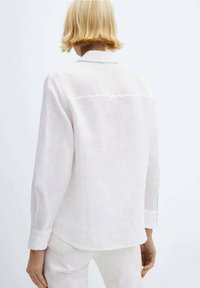 Persona con cabello rubio corto que lleva una camisa blanca de manga larga y pantalones blancos, vista desde la espalda sobre un fondo liso.