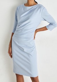 Vestido azul claro con mangas tres cuartos, escote barco y detalles drapeados en la cintura, hecho de una tela suave. Silueta ajustada.