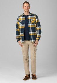 Karierte Flanellhemden in Dunkelblau, Gelb und Weiß, mit zwei Brusttaschen; kombiniert mit khakifarbenen Cargo-Hosen und braunen Schnürboots.