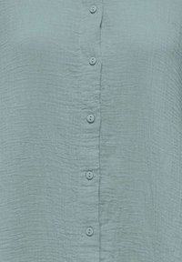 Gros plan sur un tissu vert clair texturé montrant une rangée verticale de six boutons ronds assortis.