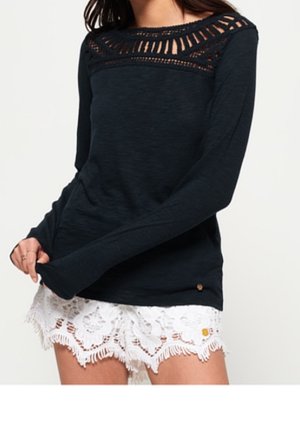 Femme portant un haut noir à manches longues avec des détails en dentelle au niveau de l'encolure et un short blanc en dentelle avec un ourlet à franges.