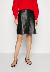 Personne portant un pull rouge, une jupe trapèze en cuir noir avec poches avant, et des sandales à talons noires à brides, debout sur un sol gris.