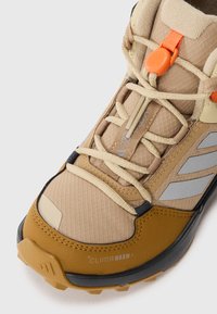 Béžová outdoor obuv s priedušnou sieťovinou a syntetickými materiálmi, gumovou podrážkou, reflexnými akcentmi a nastaviteľným oranžovým šnurovacím systémom.