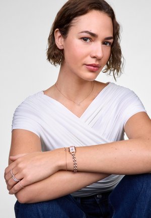 Montre en or rose avec un cadran carré, accompagnée de bracelets délicats et de boucles d'oreilles en forme de cœur, sur un haut blanc et un jean bleu.