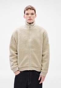 Bež flis jakna s visokim ovratnikom, punim prednjim patentnim zatvaračem, rebrastim manšetama i donjim rubom. Ima teksturirano tkanje i opušteni kroj.