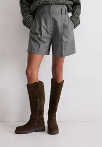 Shorts en laine grise avec des plis, associés à des bottes en daim marron foncé arrivant jusqu'aux genoux, dotées d'une fermeture éclair sur le côté et d'une semelle épaisse. Un pull en tricot texturé est visible.