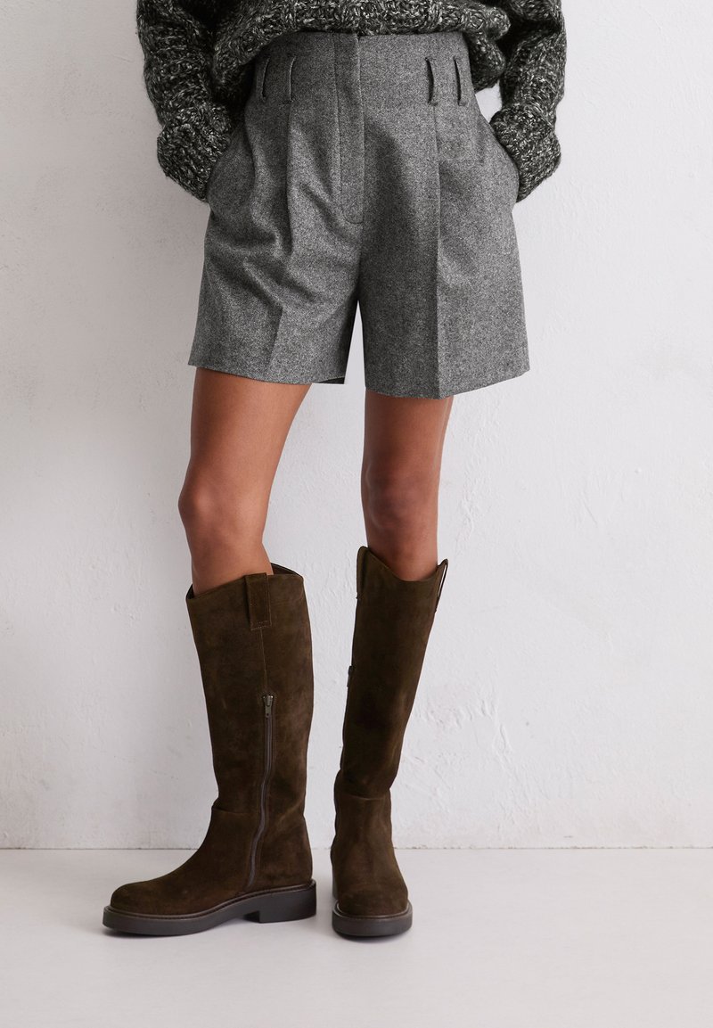 Shorts en laine grise avec des plis, associés à des bottes en daim marron foncé arrivant jusqu'aux genoux, dotées d'une fermeture éclair sur le côté et d'une semelle épaisse. Un pull en tricot texturé est visible.