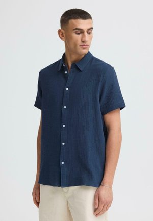 Mann in einem marineblauen kurzärmeligen Hemd mit Knöpfen und hellbeigen Hosen, der nach unten und zur Seite schaut vor einem einfarbigen Hintergrund.