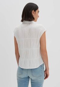 Blusa bianca a maniche corte con strisce verticali testurizzate, orlo curvo e colletto, abbinata a jeans azzurri chiari.