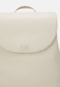 Beżowy plecak wykonany z teksturowanego materiału, z klapką zapinaną na zatrzask, tłoczonym logo CK oraz subtelnym powtarzającym się wzorem nazwy marki.