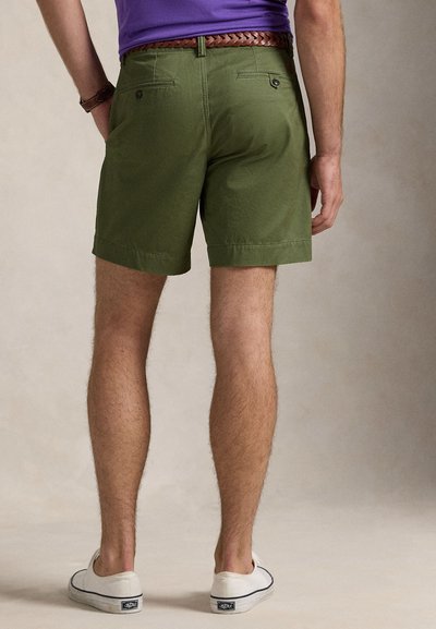 Olivovo zelené šortky z bavlny s rovným predným dielom, dvoma zadnými vreckami a zrolovanými lemami, v kombinácii s bielymi plátenými topánkami.