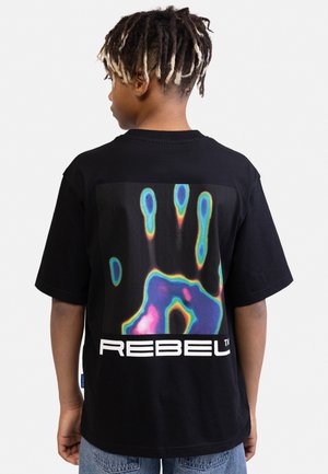 Ung person med korte dreadlocks har på seg svart t-skjorte med fargerikt varmekart-håndavtrykk og teksten "REBEL" på baksiden.
