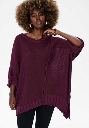 Vrouw met krullend haar, gekleed in een losse, oversized paarse gebreide trui en zwarte broek, poseert met een gebogen arm en haar hand dicht bij haar nek.