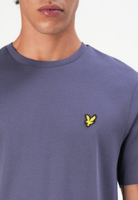 Hombre que lleva una camiseta de cuello redondo morada con un pequeño emblema de un pájaro amarillo en el lado izquierdo del pecho, cortada a la altura del cuello y a media torso.