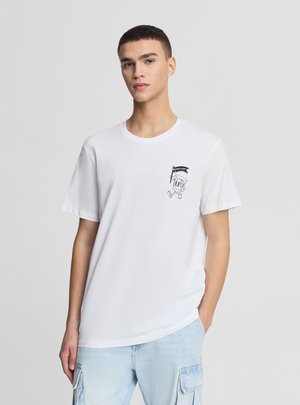 Giovane uomo con maglietta bianca con piccolo personaggio dei cartoni stampato che tiene una bandiera, abbinata a pantaloni cargo azzurri.