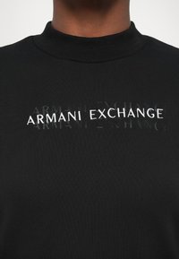 Čierny sveter s vysokým golierom, s bielym logom "ARMANI EXCHANGE". Hladký povrch a čiastočne vystúpené písmená.
