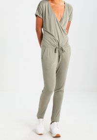 Une combinaison vert olive en tissu doux et léger, avec un décolleté en V profond, des manches courtes, une taille cintrée à nouer et des jambes fuselées.