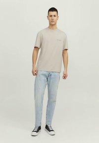 Mladý muž stojící, oblečený v béžovém tričku s krátkým rukávem, světle modrých džínách a černých vysokých teniskách s bílými tkaničkami.