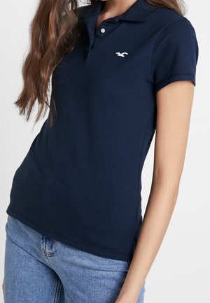 Woman wearing a fitted navy blue polo shirt with a small white logo on the chest and light blue jeans, standing against a plain background.