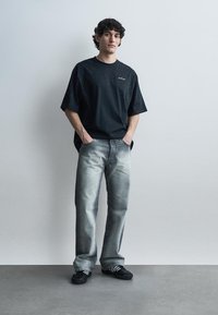 Giovane uomo con capelli ricci che indossa una maglietta nera oversize a macchie, jeans a gamba dritta sbiaditi e scarpe da ginnastica nere, in piedi contro un muro chiaro e semplice.