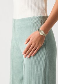 Pantaloni din corduroy verde mentă cu o textură ribbed, asortați cu un ceas argintiu cu față rotundă și accente din cristale pe un brățară din lanț.