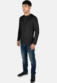 Schwarzes langärmliges Henley-Shirt mit Holzknöpfen, runder Ausschnitt, weicher Textur, kombiniert mit blauen Jeans und schwarzen Sportschuhen.