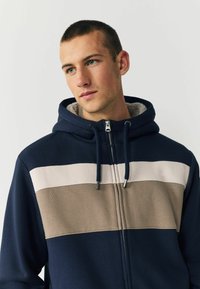 Námořnická zipová mikina s horizontálními barevnými bloky v světle béžové a taupe. Vyrobená z měkké tkaniny s fleecovou podšívkou.