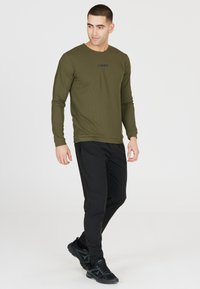 Olijfgroene lange mouw shirt met een textuurpatroon, met een zwart logo in het midden; gecombineerd met zwarte slim-fit broek en zwarte sneakers.