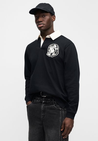 Μαύρο πόλ ο με μακριά μανίκια, λευκό γιακά και γραφικό αστροναύτη στο αριστερό στήθος. Συνδυασμένο με σκούρο τζιν και μαύρο καπέλο.