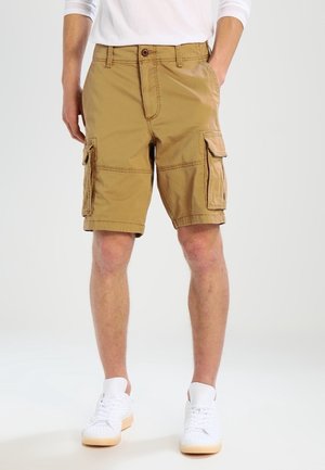Khaki Cargo-Shorts mit mehreren Seitentaschen, Knopfverschluss und glattem Baumwollstoff. Getragen mit weißen Sneakers.