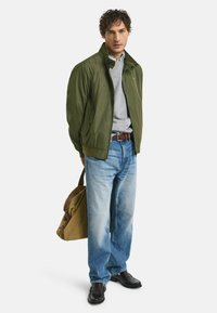 Giacca bomber verde, maglione grigio sotto, jeans azzurri chiari e scarpe nere. Cintura marrone. Porta una borsa a tracolla marrone e beige.