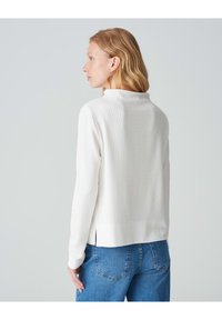 Weißer, strukturiert gestrickter Pullover mit hohem Kragen und seitlichen Schlitzen, kombiniert mit blauen Jeans. Die Farbe und das Material sorgen für einen lässigen Look.