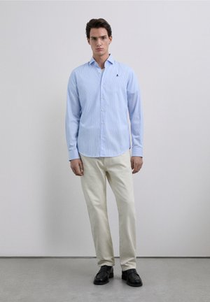 Chemise à rayures bleu clair avec col et boutons, portée avec un pantalon beige et des chaussures noires. Logo notable sur la poitrine gauche.