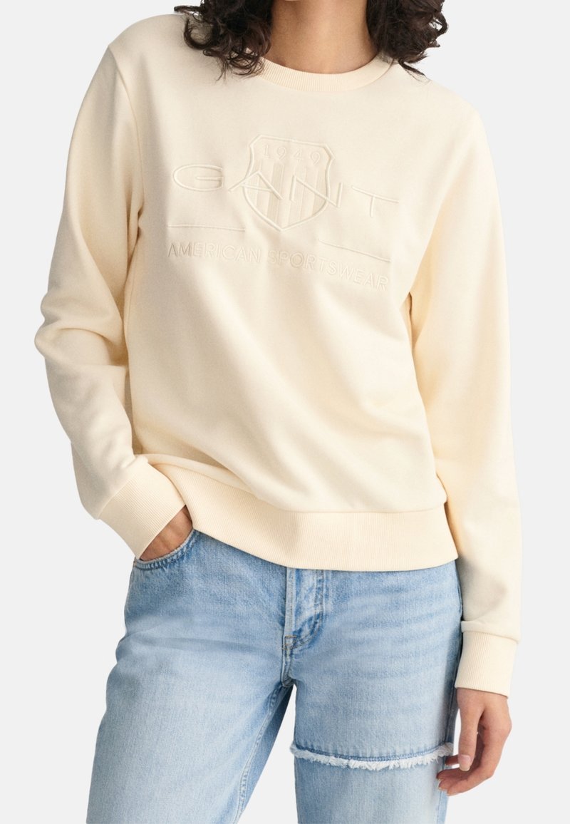 Cremefarvet sweatshirt med hævet logo; rund hals, ribbede manchetter og kant, blødt stof, parret med lys blå slidte jeans.