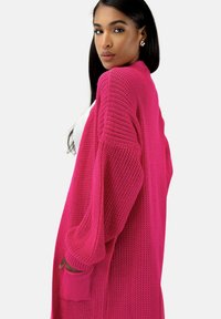 Cardigan rose tricoté à coupe ample, avec des poignets côtelés et des poches, présentant un motif texturé et un design à ouverture frontale.