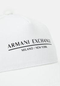 Balta cepure no gludas auduma, kurai uzrakstīts "ARMANI EXCHANGE" lieliem melniem burtiem, zem tā mazākā fontā "MILANO / NEW YORK".