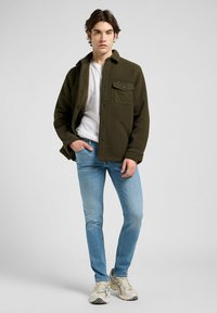 Ung mann iført olivengrønn fleecejakke, hvit t-skjorte, lyseblå jeans og beige sneakers, står med en hånd i lomma.