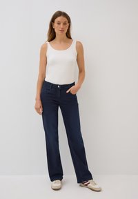 Vaqueros de mezclilla azul oscuro con corte recto, bolsillos delanteros y cierre de botón. Combinados con una camiseta sin mangas blanca y zapatillas blancas.